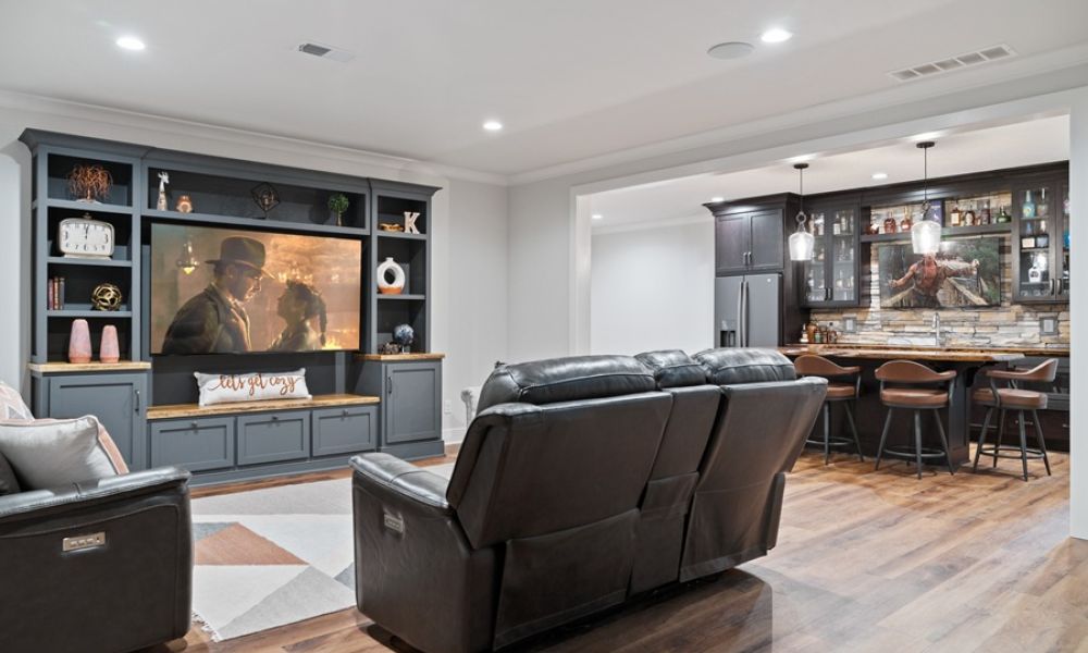 <span class="uk-h4">Basement media room with a gray built-in entertainment center, leather reclining sofa, and a wet bar with barstools.</span>