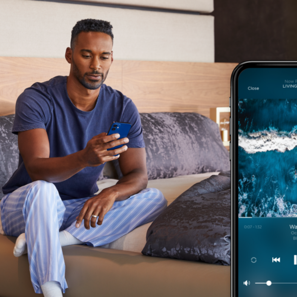 <span class="uk-h4">Man relaxing in the bedroom while controlling whole-home audio on a smartphone app</span>