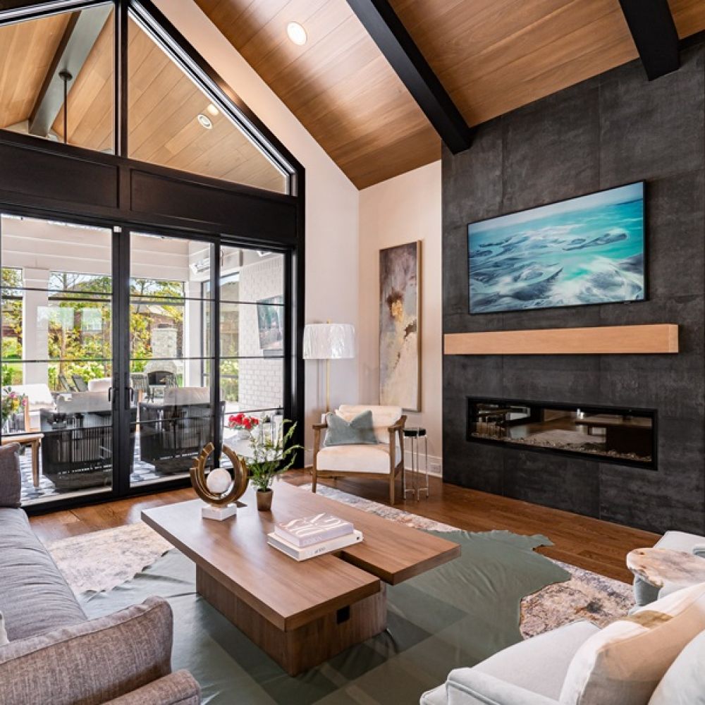 <span class="uk-h4">Modern living room with a wall-mounted TV over a black fireplace, vaulted wood ceilings, and large glass patio doors.</span>