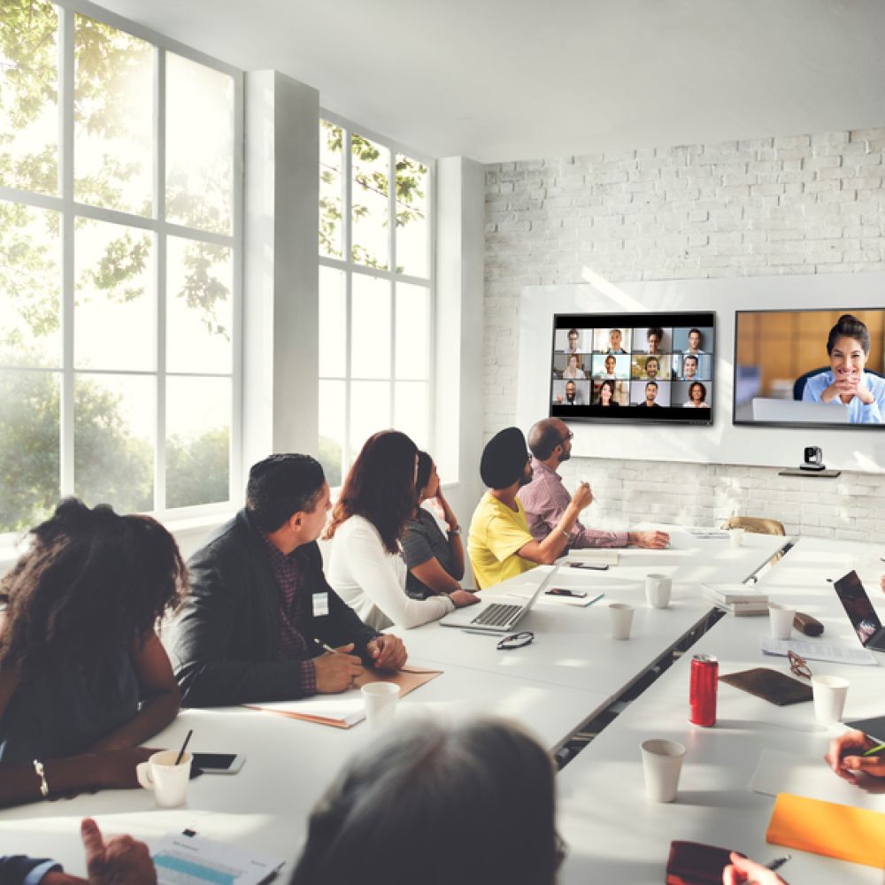 <span class="uk-h4">A large conference room with a long table, multiple people seated, and three screens in the front for videoconferencing.</span>