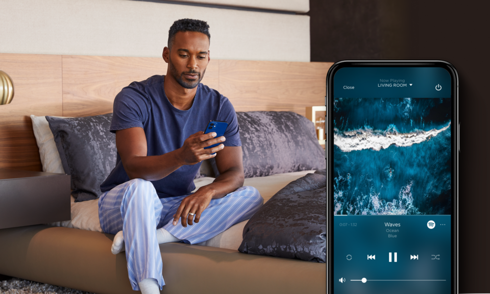 <span class="uk-h4">Man relaxing in the bedroom while controlling whole-home audio on a smartphone app</span>