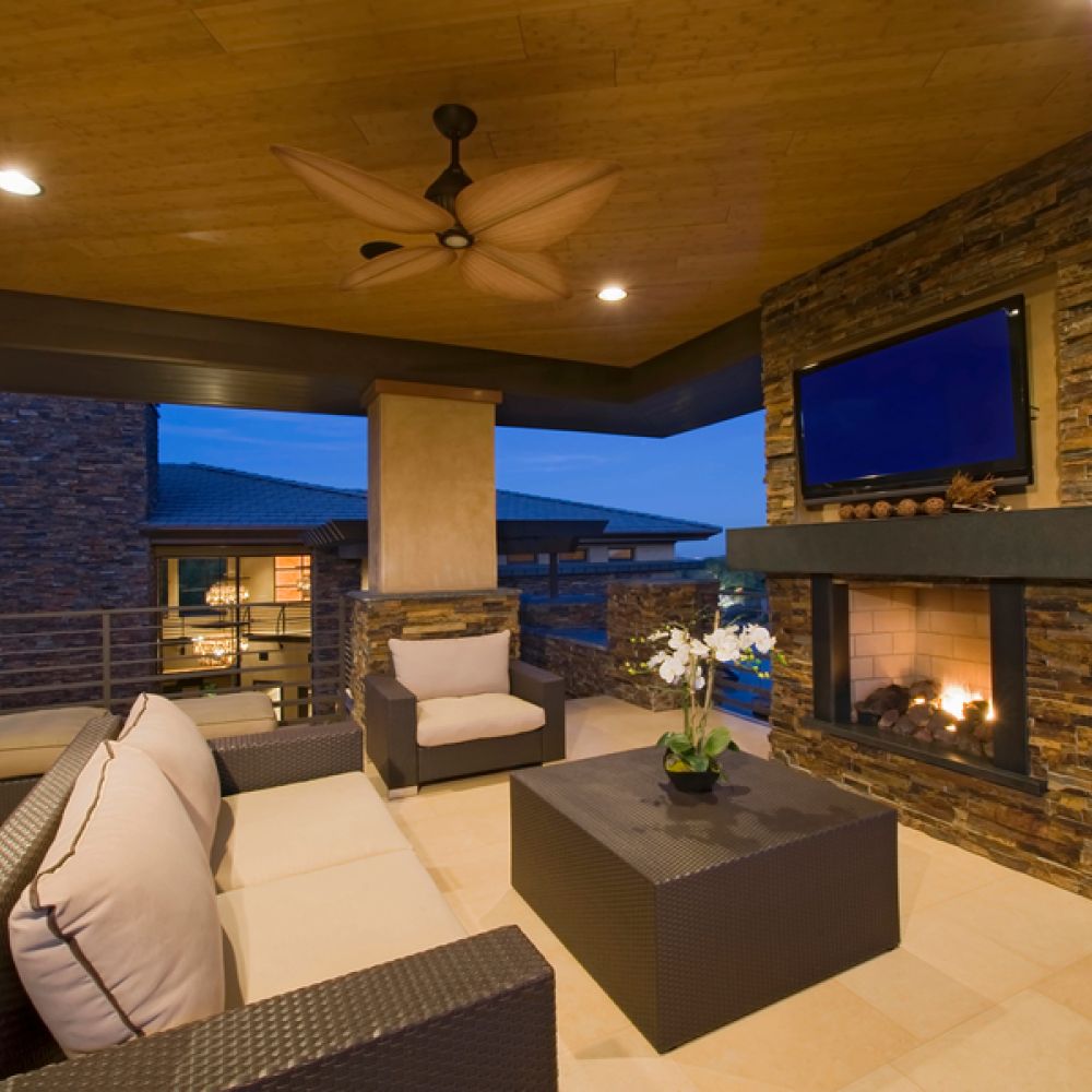 <span class="uk-h4">Modern outdoor patio with a stone fireplace and mounted TV, cushioned wicker seating, recessed ceiling lighting, and a warm, inviting atmosphere at dusk.</span>