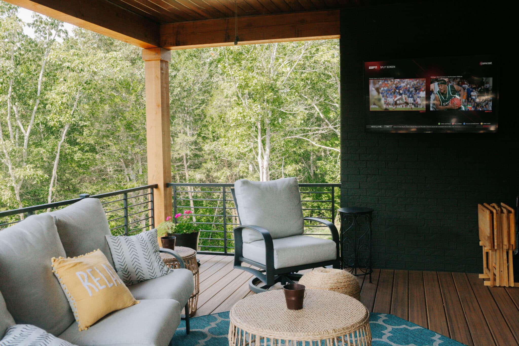 Covered patio with comfortable seating, a blue rug, and a large TV mounted on a dark brick wall.